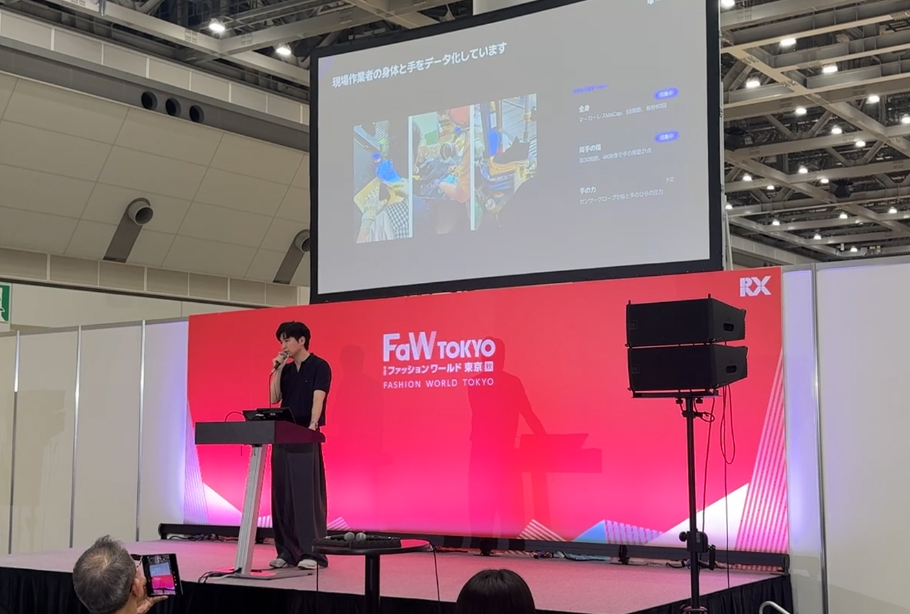 赤いFaW TOKYOのステージで発表者がマイクを持ち、上のスクリーンに工場作業と動作データの資料が映っています。
