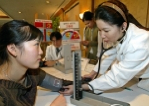 목표혈압 140/90mmHg 미만유지의 중요성을 '혈압계' 및 '자동차 속도계'로 표현한 조형물 뒤에서 젊은 여성과 남성들이 고혈압 가능성을 점검 받고 있는 모습