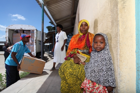 어머니 나스라 알리(Nasra Ally)와 아이가 탄자니아의 다르 에스 살람 지역에 있는  병원 밖에서 트럭이 의약품을 직접 가져다 놓는 것을 기다리고 있다.