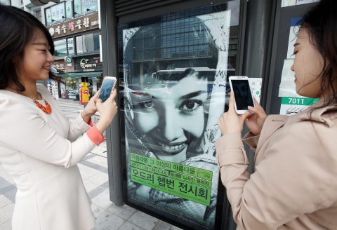 버스 정류장에서 버스를 기다리는 사람들이 오드리 헵번, 뷰티 비욘드 뷰티 전시회 버스 쉘터 광고 속 오드리 헵번 사진을 찍는 모습