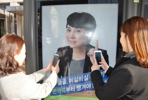 서울 신논현역 버스 정류장에서 버스를 기다리는 사람들이 내츄럴엔도텍 웰뮨 광고 인증샷 이벤트에 참여하기 위해 광고 사진을 찍는 모습